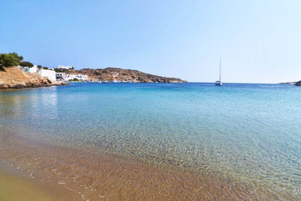 A low view of a sandy beach in Sifnos with clear turquoise water. There is a small mountain with white buildings in the background and one sailboat in the water.