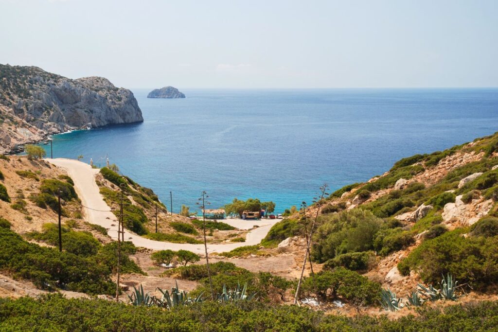 A panoramic view of Vroulidia, an off-the-beaten-path Sifnos beach. The blue water is surrounded by lush green plants and a gravel road leading down to the beach. You can see cliffs in the distance on the left.