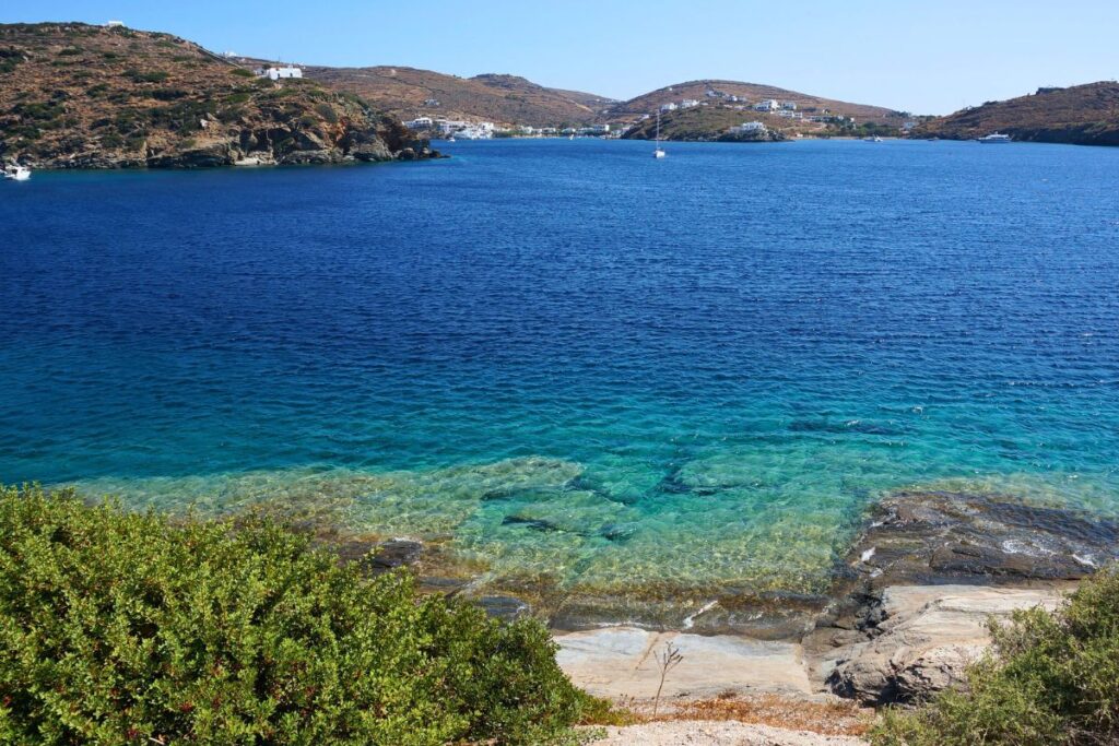 A beautiful sea view from Vroulidia of the stunning blue Aegean Sea. There is a rocky ledge and green plants in the foreground and small mountains in the distance.