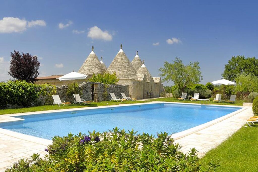 The beautiful pool at the Abate Masseria hotel in Alberobello, Puglia. There are white loungers and umbrellas surrounding the pool and traditional trulli in the distance.