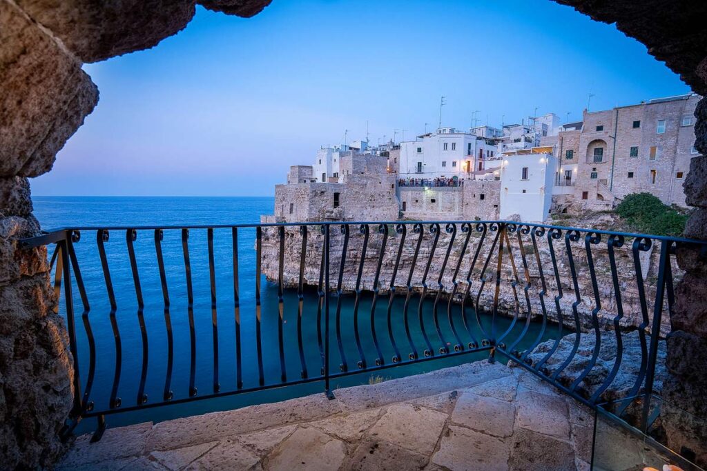 A stunning view of Polignano a Mare, one of the most beautiful towns in Puglia to visit. The town sits on limestone cliffs that tumble into the ocean and the view is from a private patio with a rock wall at dusk.