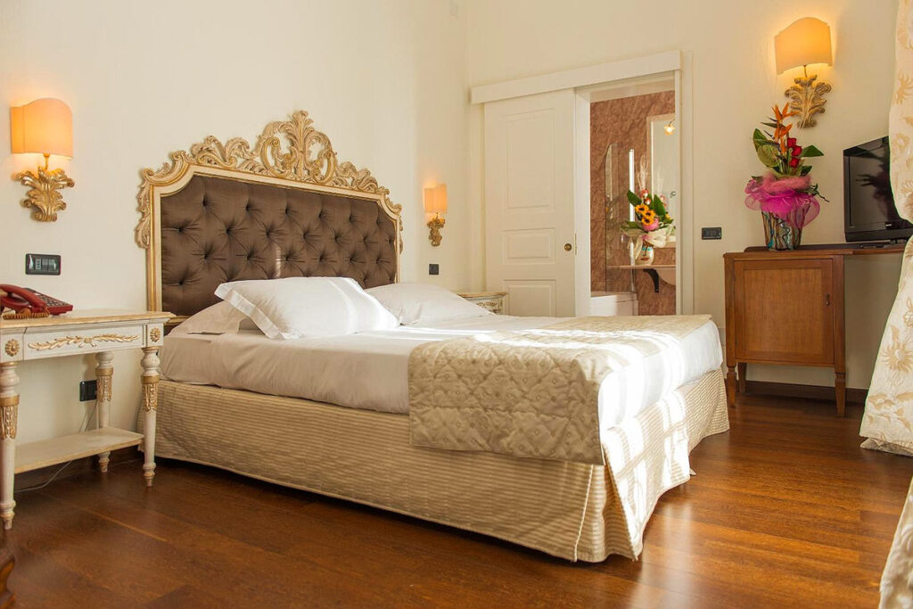 A classically designed room at the Grand Hotel di Lecce. There is a queen bed with a regal, elegant headboard, and a door leading to a spacious, marble bathroom.