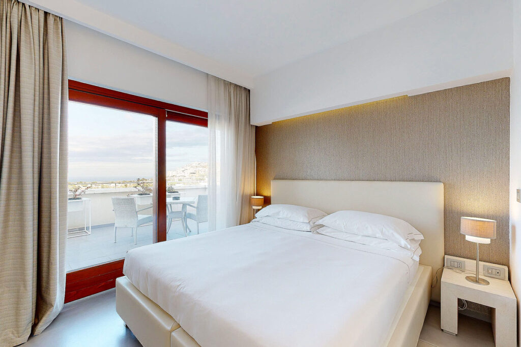 A clean, modern room at Monte Sarago hotel with a white comfortable bed, two side tables and sliding doors that lead out to a huge patio. You can see Ostuni in the distance, one of the best towns in Puglia to visit.