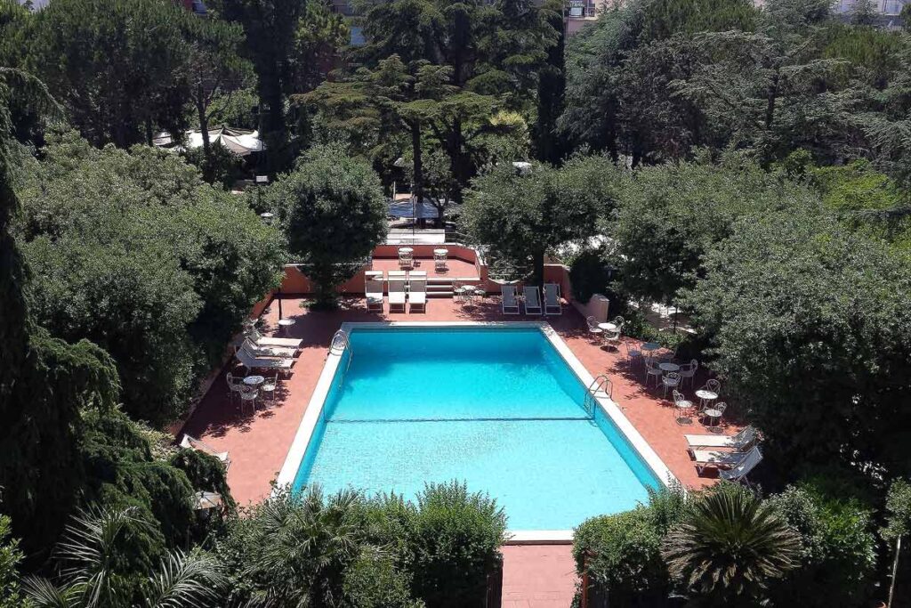 A bird's eye view of the pool at the Park Hotel San Michele in Martina Franca. Red concrete surrounds the pool and there are white loungers spread about and lots of huge green trees.