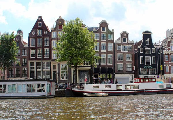 A row of crooked houses on a canal in Amsterdam. There is one single green tree to the left of the center with a large boat docked on the right.
