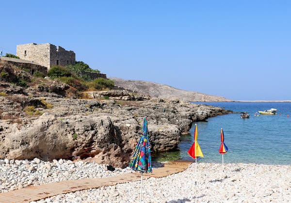 A white pebbled beach in Greece with a wooden ramp leading into the blue sea. There are 3 colorful umbrellas on the right and a rocky coastline on the left with an old stone building.