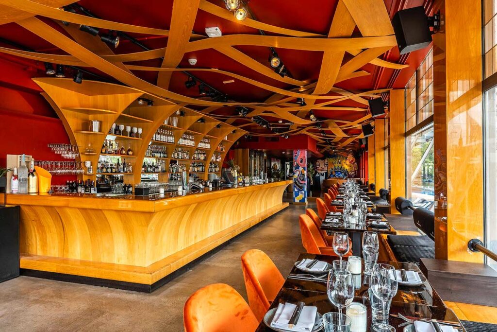The vibrant eclectic bar at the Apollo Hotel Amsterdam, a Tribute Portfolio Hotel (Marriott). The ceiling is an abstract design of red and gold fabric with a wood paneled bar underneath. There are tables and chairs on the right overlooking the canal.