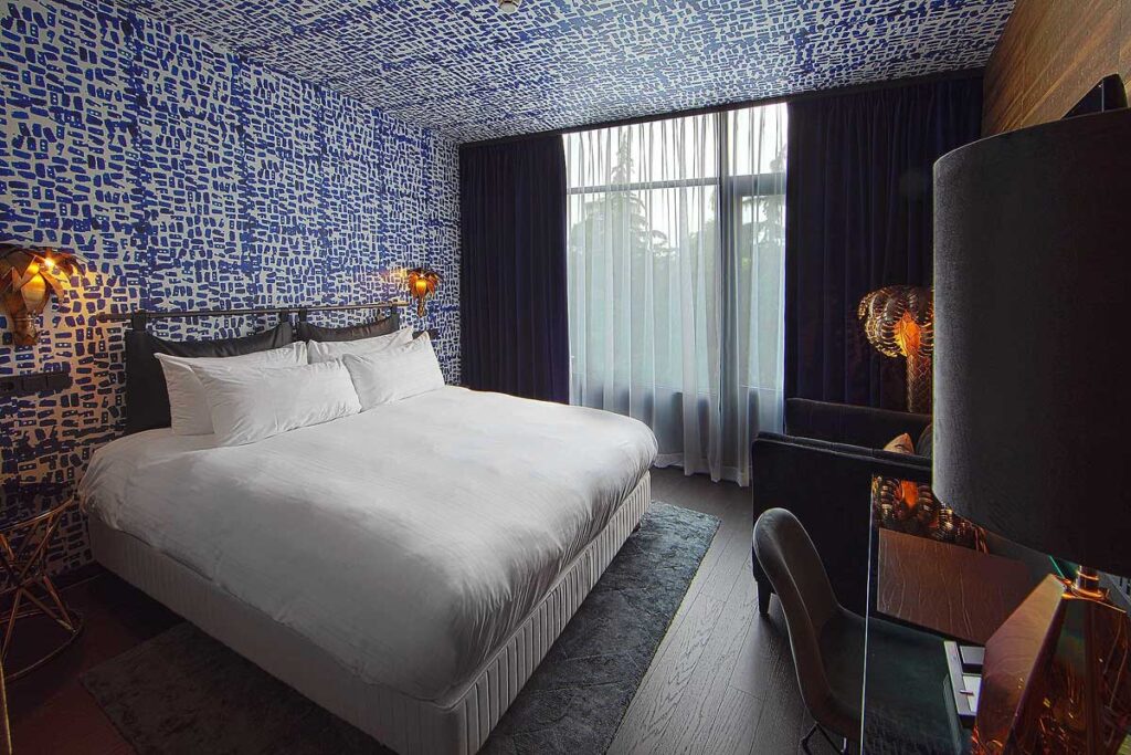 A stunningly designed room at the Apollo Hotel Amsterdam, one of the best wheelchair accessible hotels to stay at. There is blue and white abstract wallpaper on the back wall that runs up the ceiling with a full length window and balcony door at the back. On the left is a large white bed with a small desk and chair on the right.