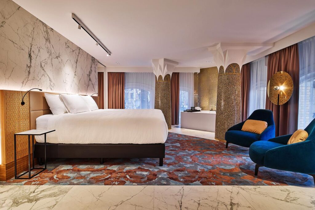 A spacious and beautifully designed room at the Clayton Hotel Amsterdam American, an accessible option. There is a large white bed on the left with two bright blue chairs opposite sitting on a floral carpet. There is a deep soak tub at the back with two decorative columns around.