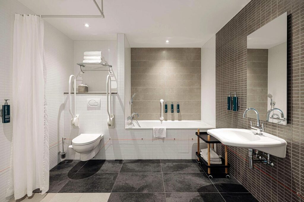 A large, spacious bathroom at the DoubleTree by Hilton Hotel Amsterdam Centraal Station, a popular wheelchair accessible hotels. There is a toilet with grab bars on the back left, and a roll under sink on the right side.
