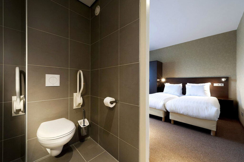 A toilet with grab bars in an espresso colored tiled bathroom. You can see through the door to the two twin beds pushed together in the spacious room at the Hotel Espresso City Centre in Amsterdam.