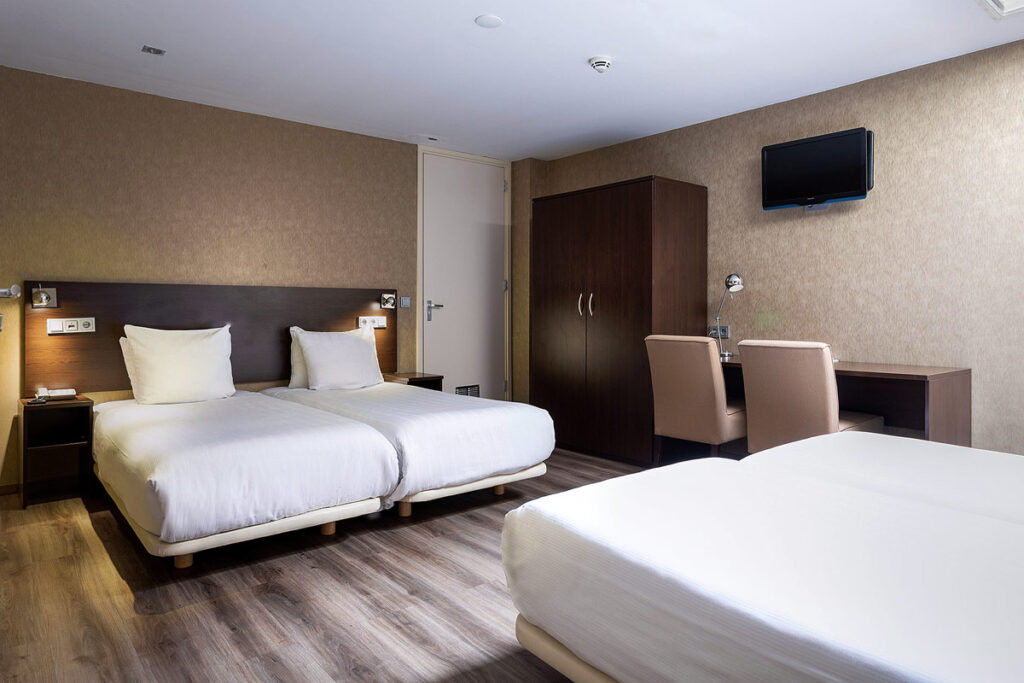 A simple yet spacious room at the Hotel Espresso City Center, a wheelchair friendly hotel in Amsterdam. The hardwood floors and walls are espresso in color and there are 3 single beds in the room, a dresser on the right and a table with 2 chairs.