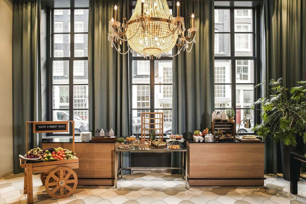 The dining room at the Radisson Blu Hotel, a wheelchair accessible hotel in Amsterdam. The room is elegant with floor to ceiling windows and sage green long curtains. There is a long wooden buffet table set up with breakfast and a cheese cart on the left side.