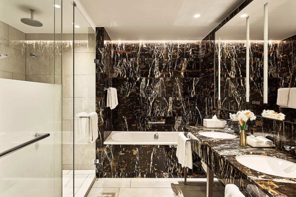 A beautifully elegant bathroom at the Hyatt Regency Hotel in Amsterdam. There is black marble tile on the far wall and over the bathtub at the back with a roll-in shower on the left and a marble roll-under vanity on the right.