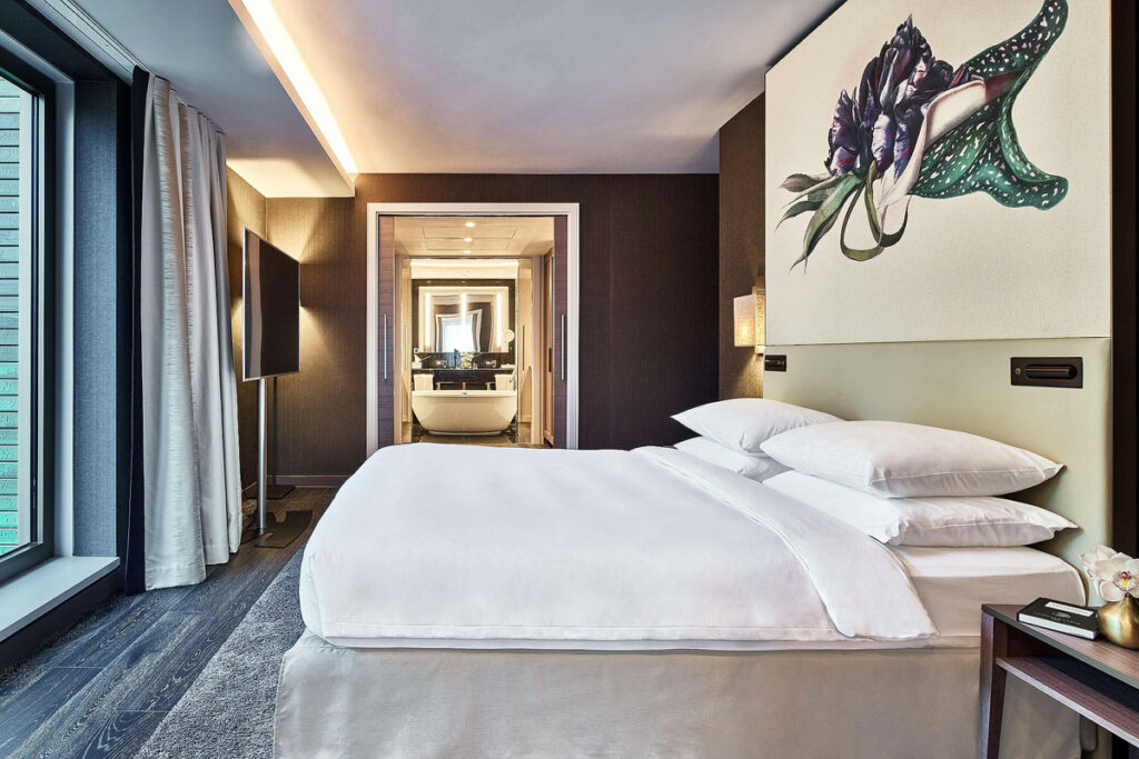 A modern room at the Hyatt Regency Hotel, a wheelchair accessible hotel in Amsterdam. The bed is on the left side with space around and a painting of a flower above. In the distance is a brown wall and a door to the spacious bathroom.