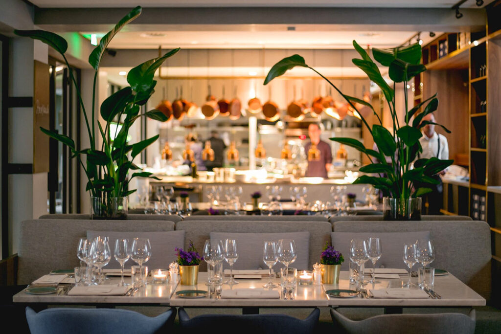 The elegant wheelchair accessible restaurant at the INK Hotel in Amsterdam. A row of tables joined together is set for dinner, and just above are green plants on either side framing the open kitchen behind with copper pots hanging from above.