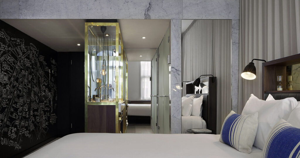 A view from above the bed to the bathroom at the INK Hotel Amsterdam – MGallery, a wheelchair friendly hotel. There are windows surrounding the vanity with gold trim and a wood counter on the left and a roll-in shower on the right.