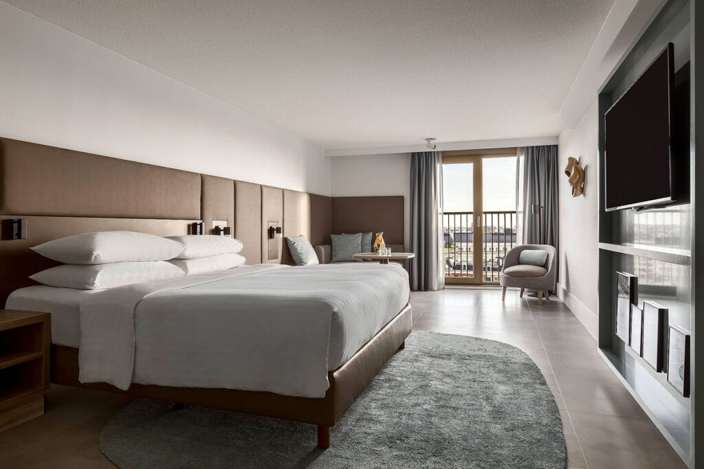 A spacious room at the Amsterdam Marriott Hotel, a wheelchair friendly place to stay. There is a large white bed on the left with wood paneling along the length of the wall, with a couple of chairs at the end beside a sliding balcony door.