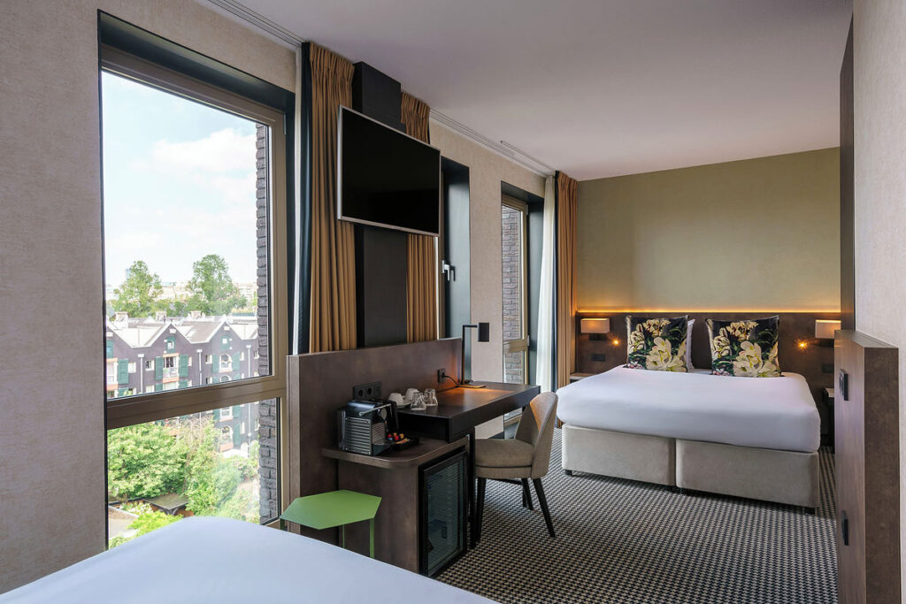 A beautiful room at the Monet Garden Hotel in Amsterdam with a view of traditional buildings through the 3 large windows. There are two large beds opposite one another and a small table with tv above in the middle.