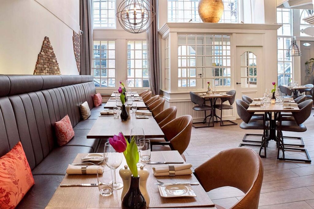 The beautiful wheelchair accessible restaurant at the Radisson Blu Hotel,
Amsterdam City Center with floor to ceiling windows at the back. There is a row of connected benches with tables and chairs on the left with random tables on the right.