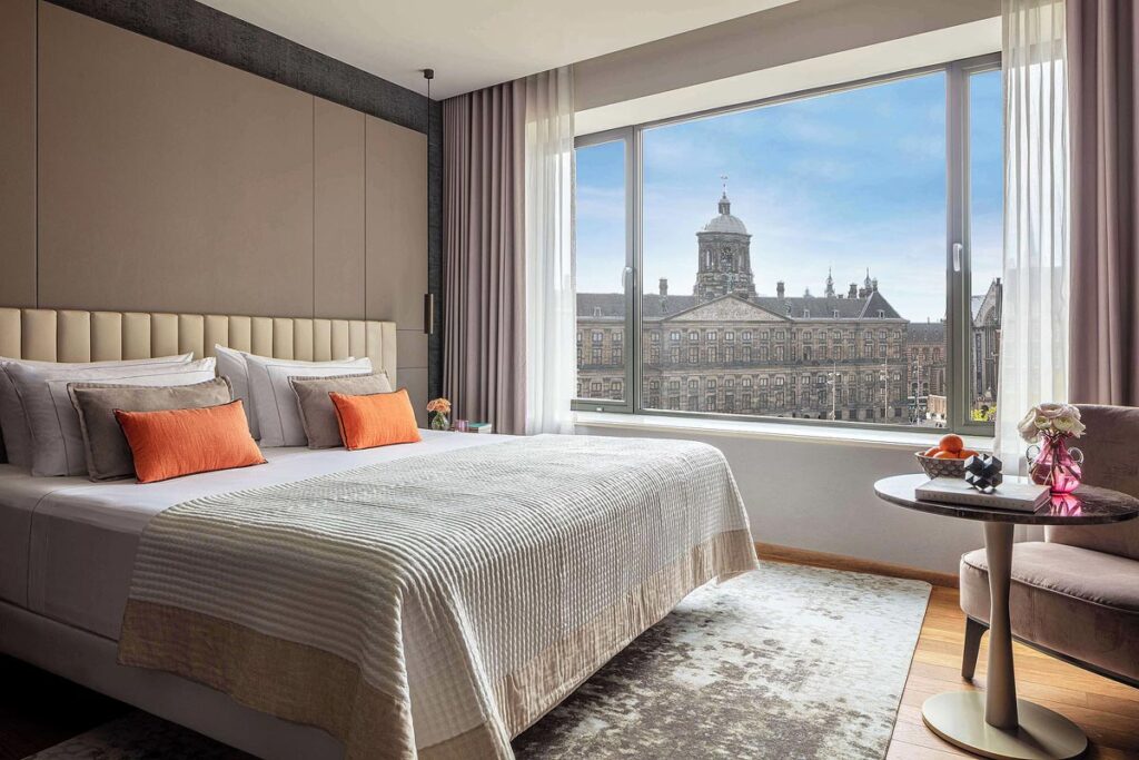 A suite in the Anantara Grand Hotel, a fantastic place to stay when visiting Amsterdam. There is a large bed with textured duvet, and grey and orange throw pillows on top of a grey textured rug. A large window overlooks the historic buildings of Amsterdam.