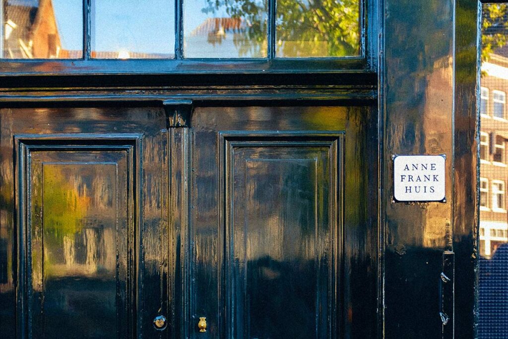 A close up of the 2 shiny deep teal door of the Anne Frank House. There is a plated sign on the right and windows above the doors reflecting the trees.