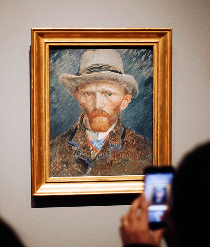 A close up of a Van Gogh self portrait of him wearing a light grey hat with black trim, and a tweed jacket with a blue background. The painting is in a large gold frame hanging on a grey wall in the museum. You can see someone taking a picture with their cell phone.
