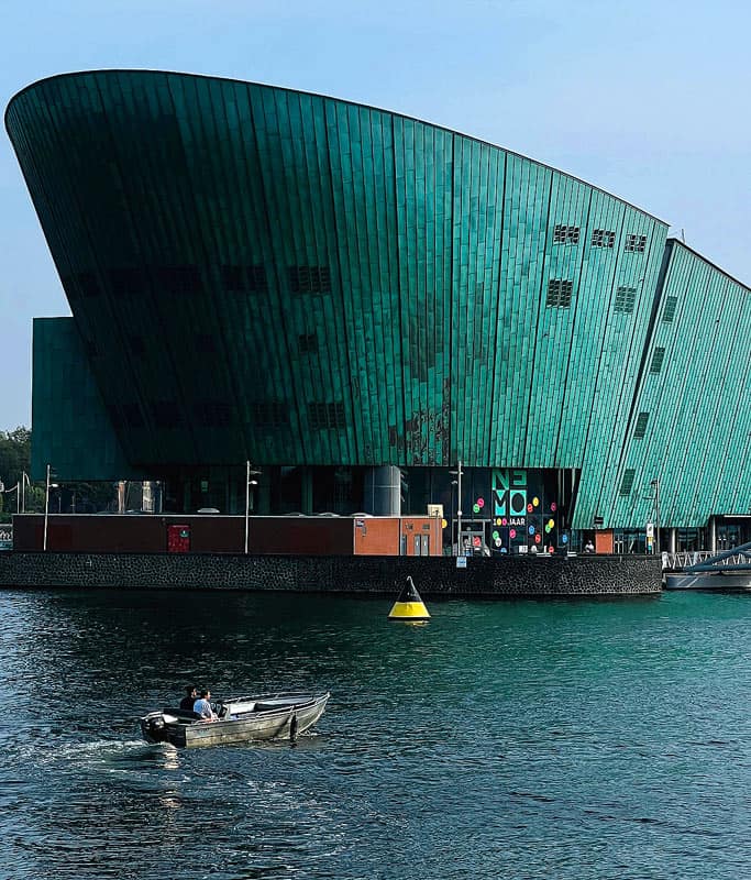 The teal green uniquely shaped building of the Nemo Science Museum, a fantastic Amsterdam tourist attraction for kids. The museum sits on the water and there is a single boat moving.