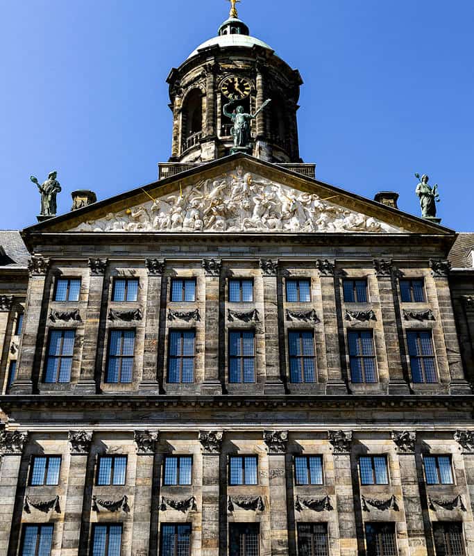 A close up of the facade of the Royal Palace, one of Amsterdam's top tourist attractions. This historic building is dark grey with rows of vertical windows and two statues on the roof.