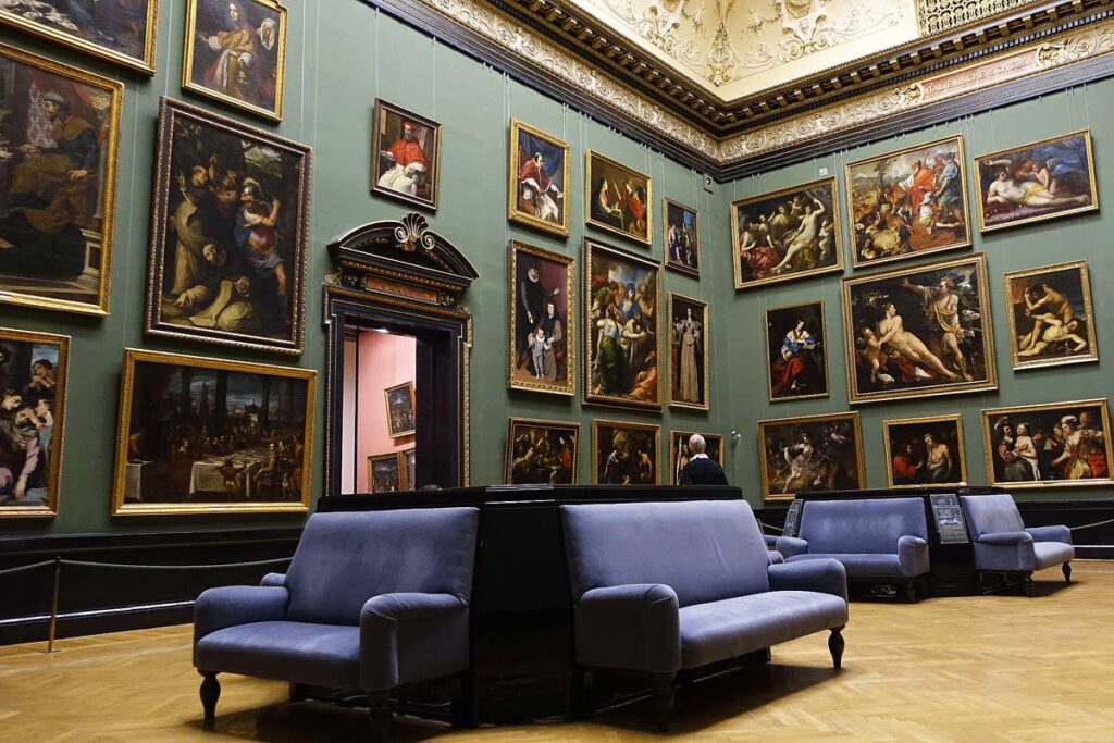 A beautiful interior room with green walls, blue couches in the center, crown moldings above, and walls full of paintings from Dutch artists at the Rijksmuseum, one of the top Amsterdam tourist attractions to visit. 