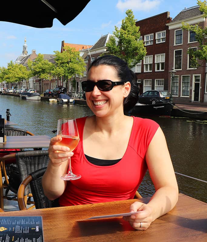 Robyn wearing a bright red shirt and sunglasses holding a glass of white wine at a table beside a canal in Amsterdam.