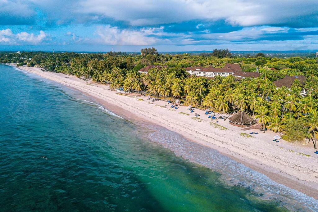 An aerial view of Diani Reef Beach Resort & Spa with the resort sitting in the tall palm trees that line the white sand and turquoise ocean under a blue cloudy sky. Photo by Expedia.
