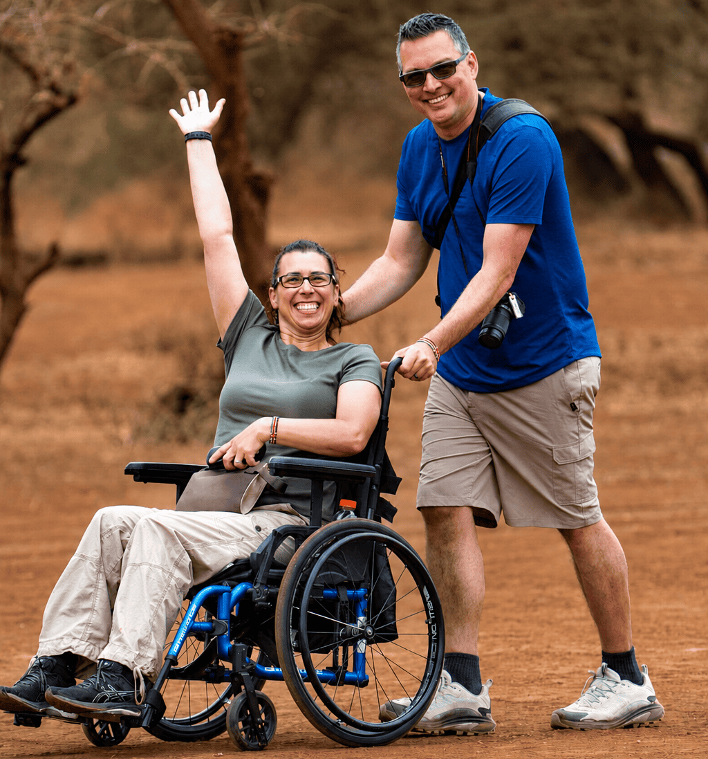 Gavin wearing a blue shirt and khaki shorts pushing Robyn in her manual wheelchair wearing all green. Both are smiling while on their accessible safari in Kenya.