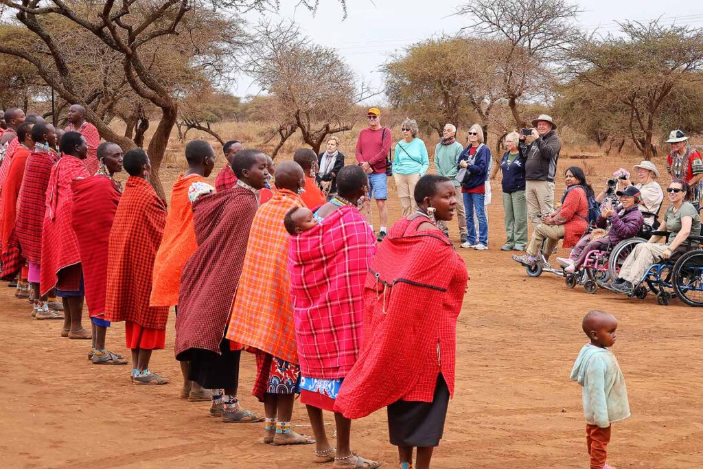 A line of Masai women and men standing and singing in front on a group of travelers, including wheelchair users. There are bare trees in the background growing on red dirt.