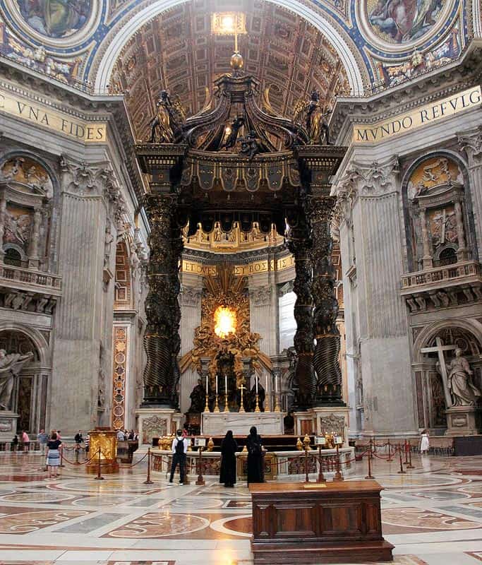 The stunning altar at St. Peter's Basilica in Vatican City, one of the top things to do in Rome as a wheelchair user. Two nuns wearing black stand in front of the massive altar with is surrounded by beautiful mosaic floors and detailed walls.