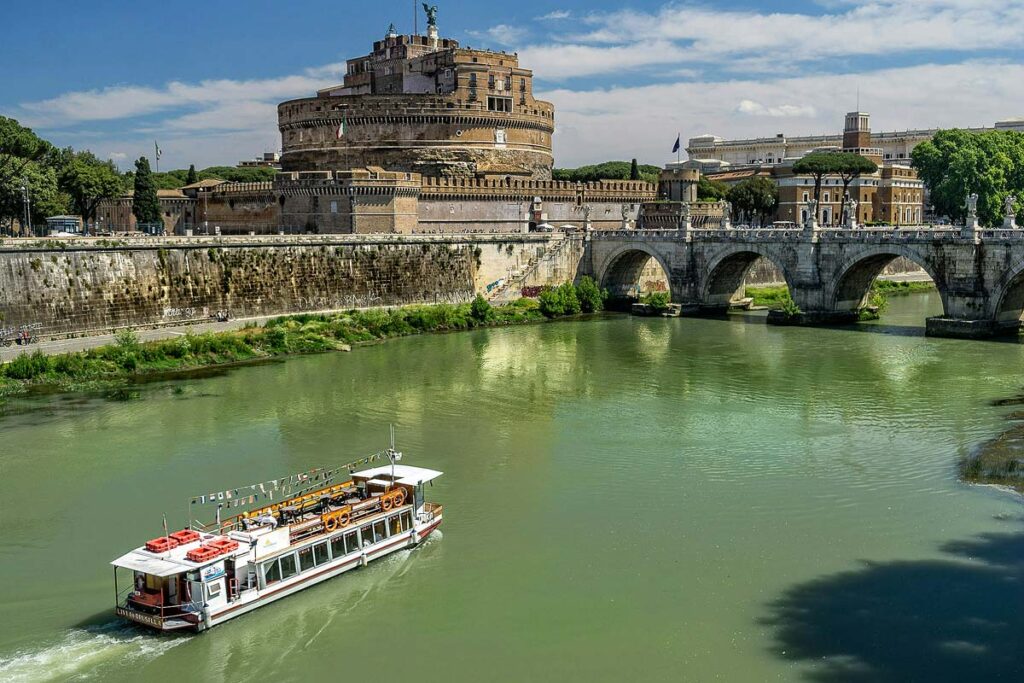 A beautiful panoramic view of Castel Sant Angelo, a highlight attraction when visiting Rome with limited mobility. The circular fortress sits along the banks of the Tiber River with a river boat sailing by.