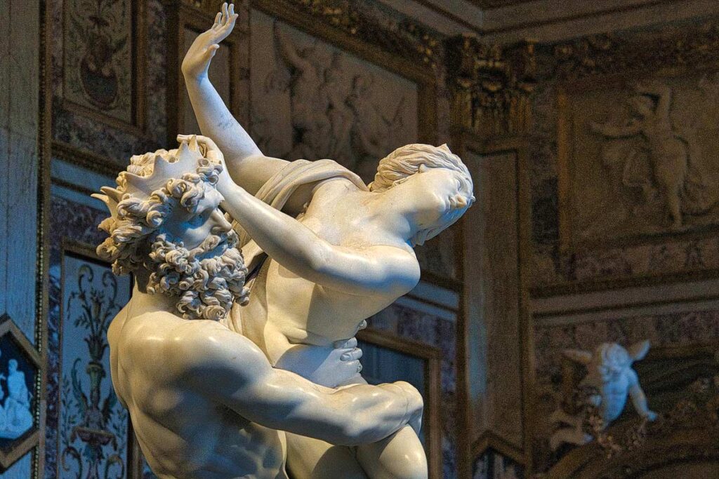 A beautiful marble sculpture of a bearded man holding a woman with highly decorated walls at the Villa Borghese Museum, a popular attraction to visit in Rome.