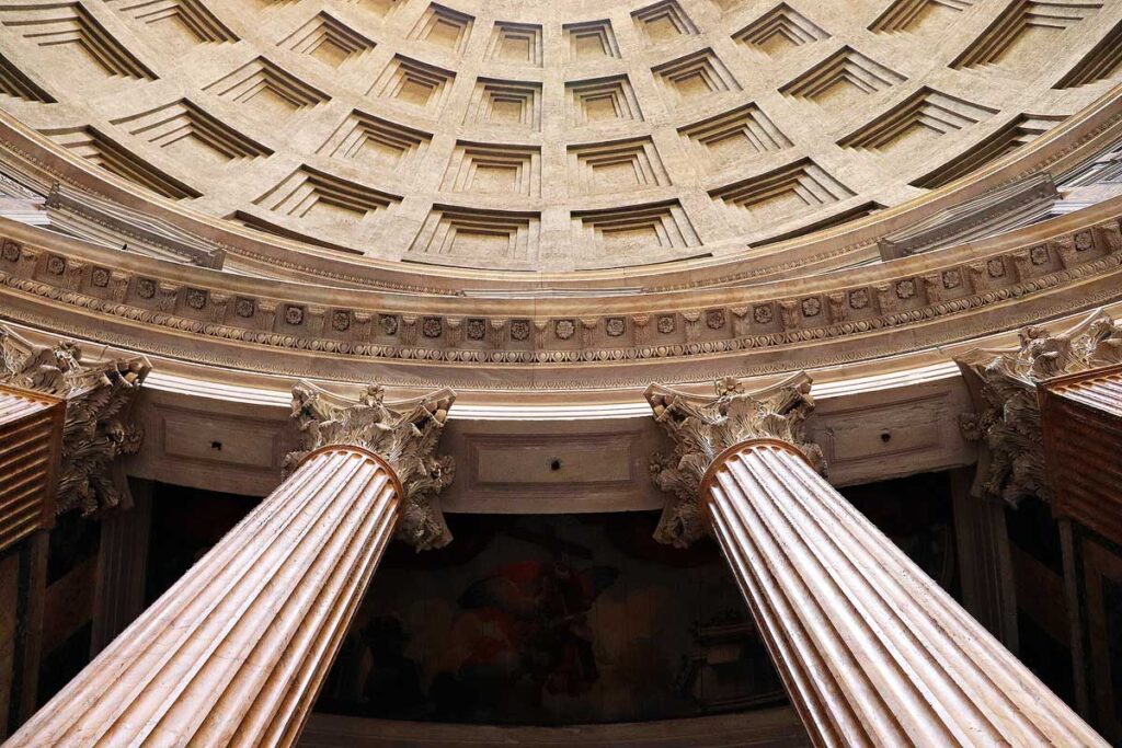 The interior arched dome of the Pantheon with two gigantic columns at the base supporting the beautifully detailed dome above, which takes up the entire frame.