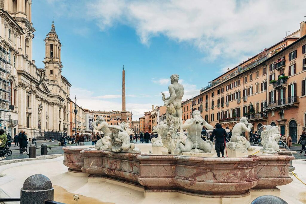 A stunning marble fountain at the center of Piazza Navona surrounded by colorful buildings and people. This is one of the top attractions to visit in Rome.