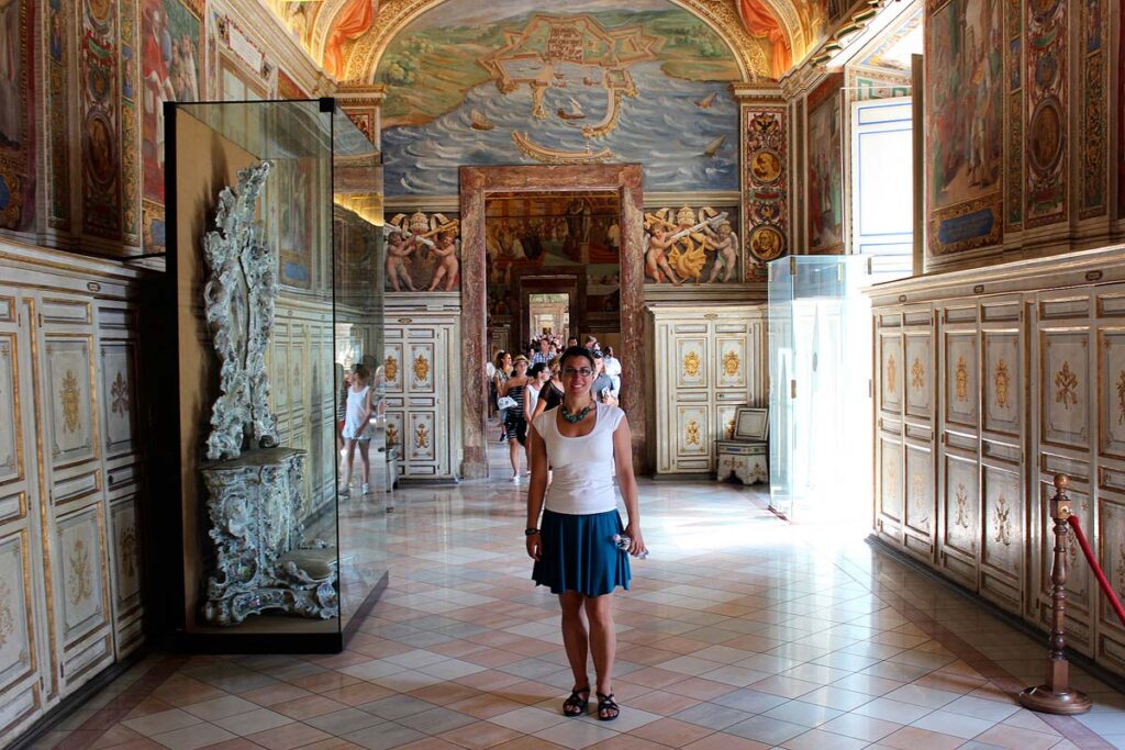 Robyn wearing a white shirt and blue skirt standing in an ornate hallway in the Vatican. The ceilings are painted with detail and there is light coming in through a window on the right.