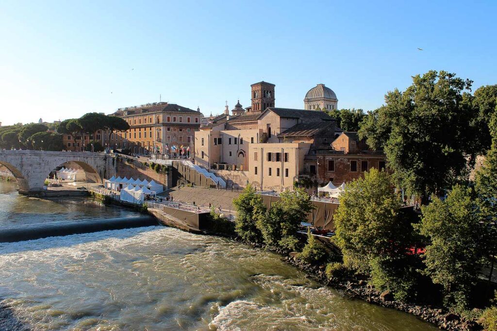 The beautiful Tiber River in Rome with peach colored buildings on the right bank with trees in the foreground and some riverside restaurants.