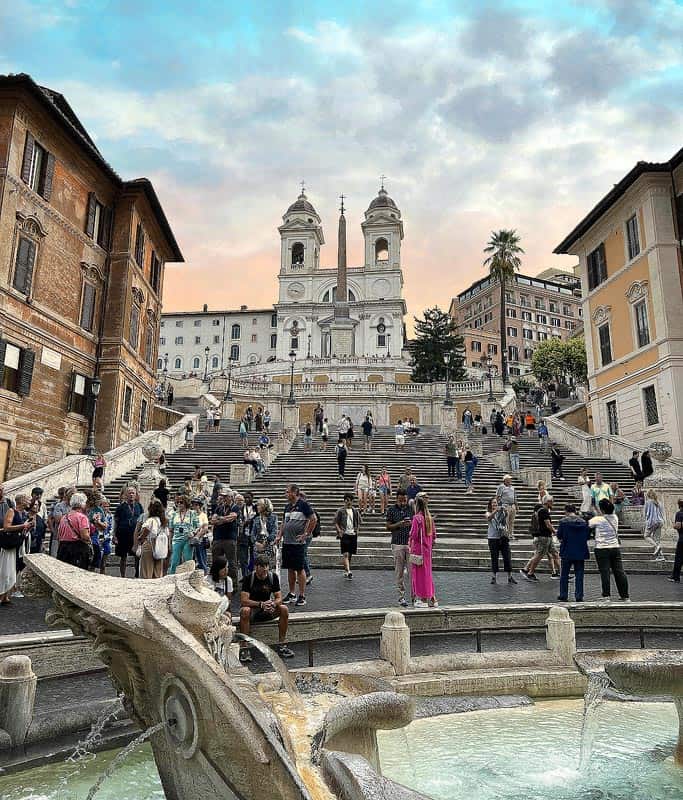 A beautiful view of the Spanish Steps from behind the fountain looking up towards the church at the top. People are around and the sky is turning from blue to pink.