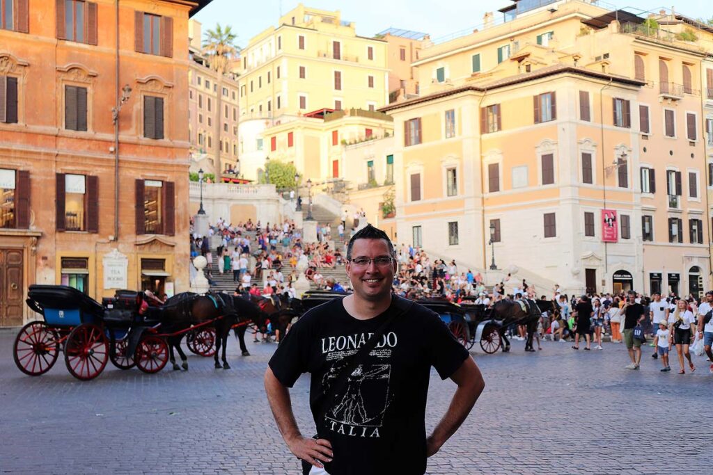 Gavin is smiling ,wearing a black shirt with the words Leonardo at the top, with the Spanish Steps in the distance full of people. There are colorful buildings around him along with a horse and carriage on the left.