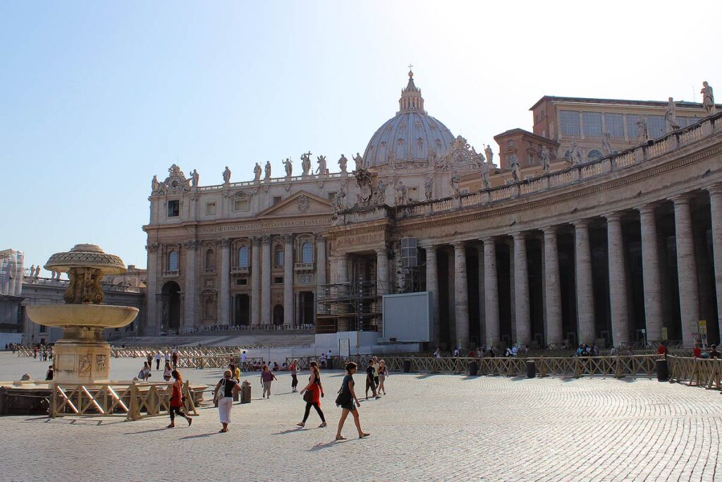 A view of St. Peter's Basilica from the right, one of the best things to do in Rome as a wheelchair user. The dome sits above center with rows of columns on the right and people walking in the square.