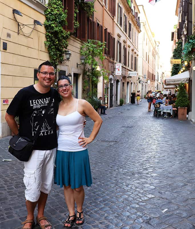 Robyn wearing a white tank top and blue skirt, standing beside Gavin in a black shirt and white shorts. They are smiling with linked arms standing to the left on a cute street in Trastevere neighborhood in Rome, a popular thing to do with limited mobility.