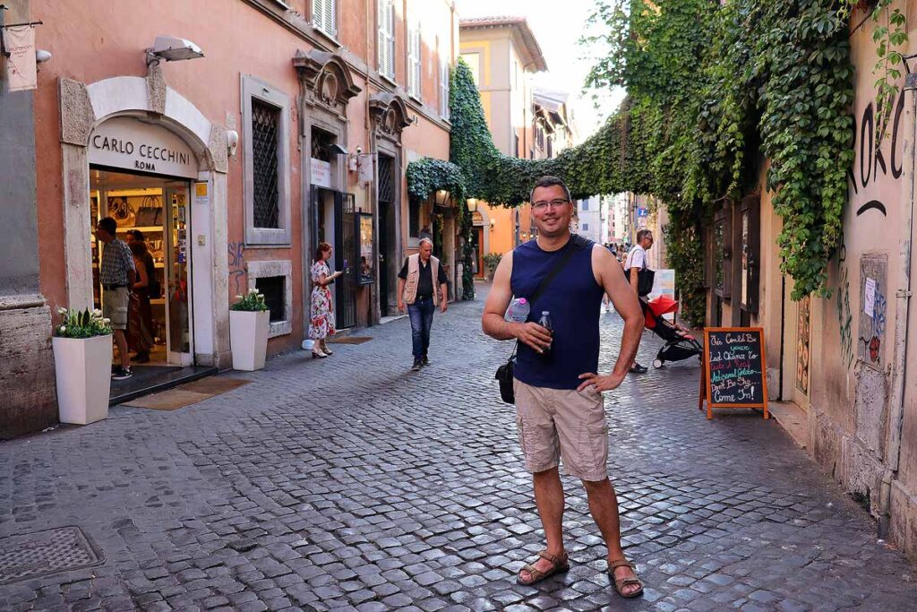 Gavin wearing a navy blue top and beige shorts on a cobbled street in Trastevere with a pink building. This neighborhood in Rome is a great place to explore with limited mobility.