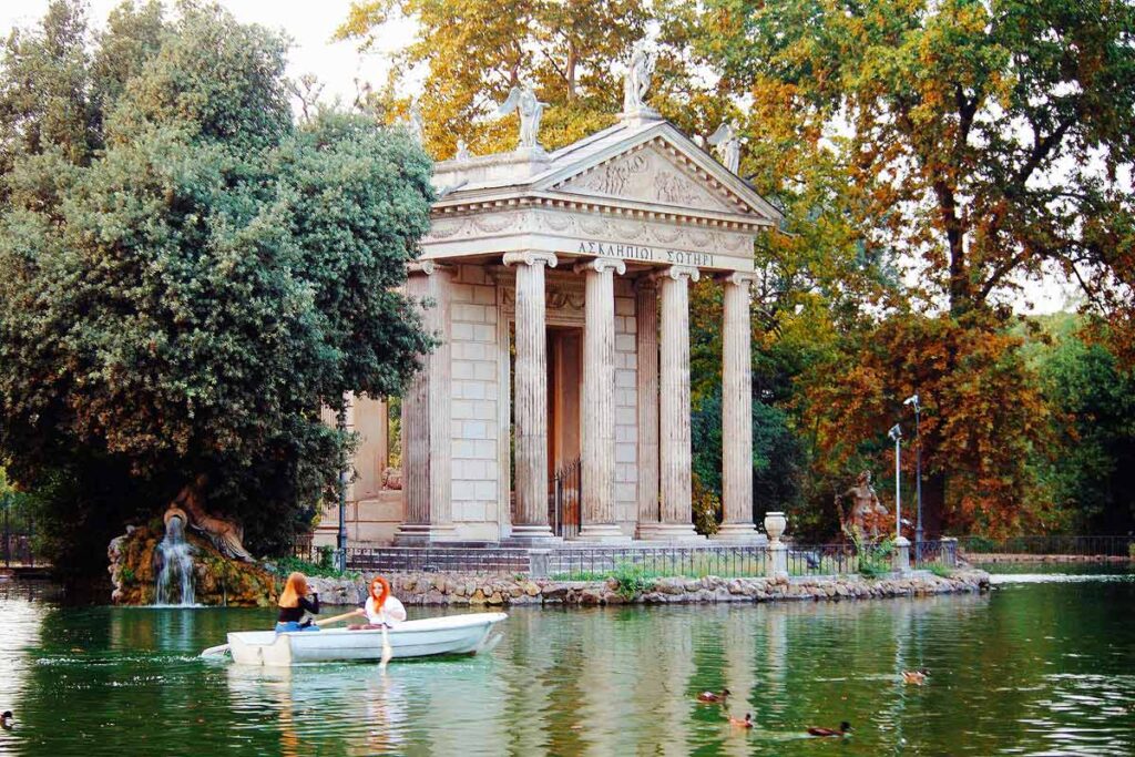 A beautiful column structure surrounded by trees sitting in front of a green lake at the Villa Villa Borghese Gardens with two people rowing in a boat.