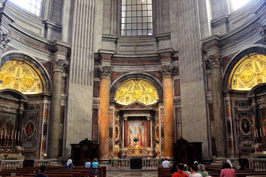 The interior of a beautiful church in Rome with pink marble columns near the altar with a woman praying, and others sitting in benches.