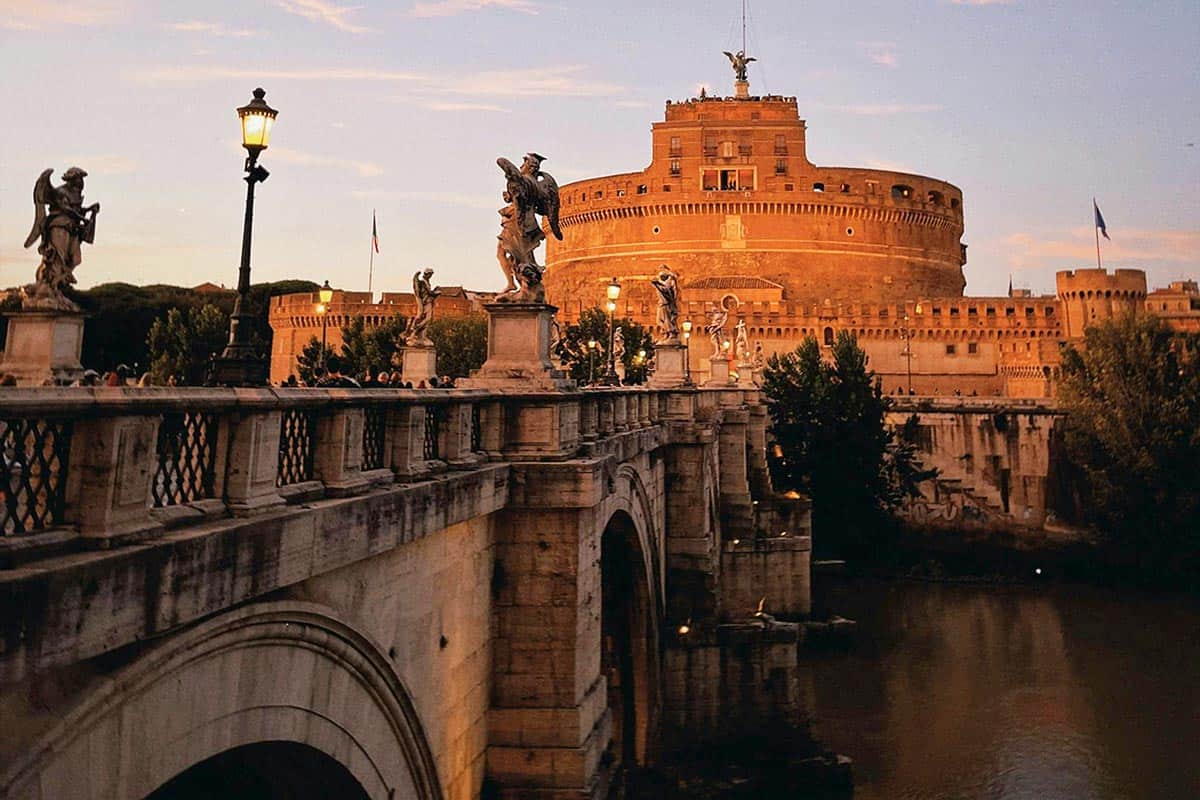 A side view of Castel Sant Angelo at sunset with golden light hitting the building and the beautiful arched bridge with lamp posts above the Tiber River.