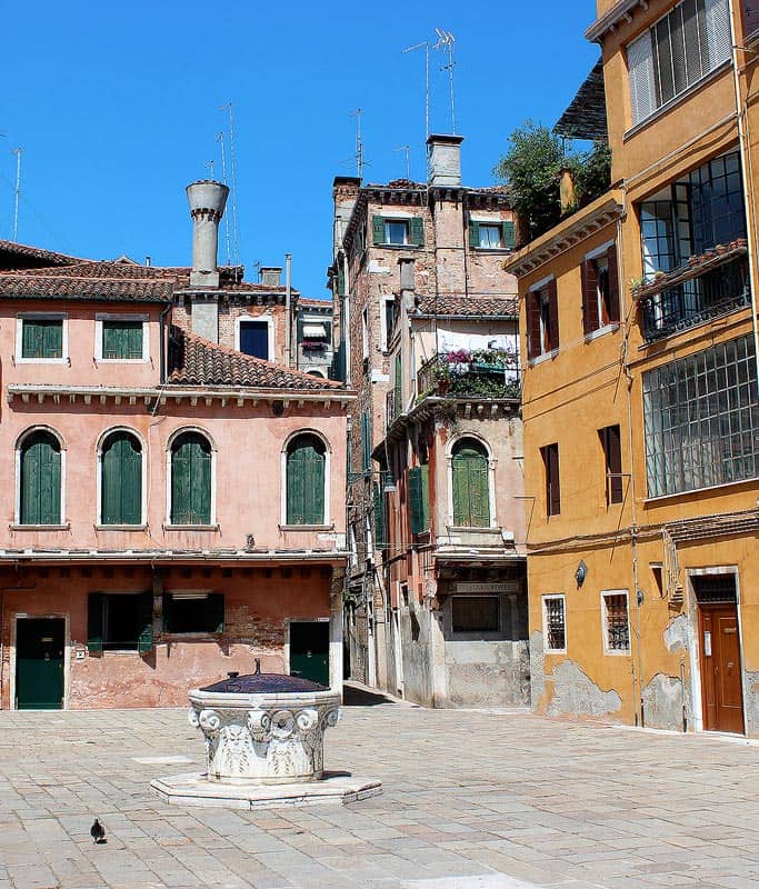 A colorful piazza in Venice, Italy. There is a pink 3 story house with green shutters on the left, and an orange 3story house with wood shutters on the right with a tiny fountain in the center.
